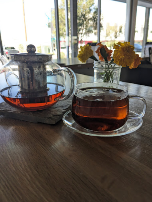 A glass teapot and cup of tea on a wooden table, with a vase of flowers in the background.